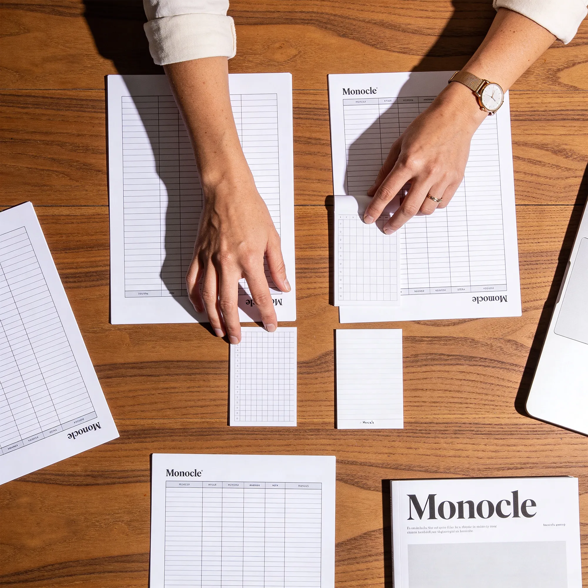 Two hands organizing Monocle-branded planner sheets and note cards on a wooden desk with a laptop partly visible.